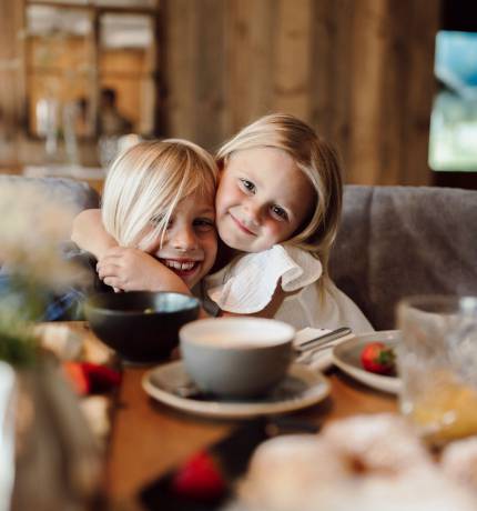 Mädchen umarmt lachenden Jungen am gedeckten Tisch im Restaurant Glückskind Ferienresort Central Flachau