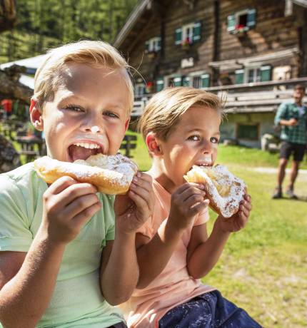 Zwei Kinder essen Krapfen auf einer Bank vor einer Berghütte