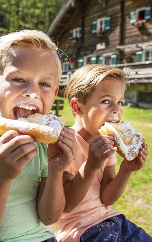 Zwei Kinder essen Krapfen auf einer Bank vor einer Berghütte